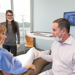 Dr Michael Stewart with female patient getting ready to give her dental work, assistant in the background aims lighting at Stewart & Arango Oral Surgery